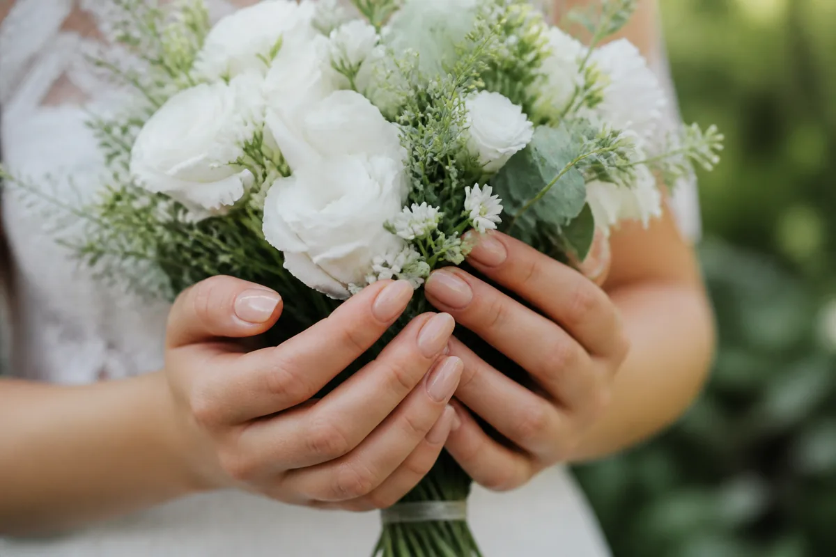 Mains de mariée aux ongles en amande courte posées sur un bouquet blanc, lumière naturelle douce